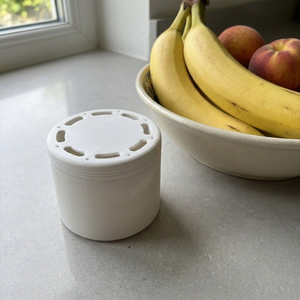 A small white lidded fruit fly trap on a kitchen windowsill next to a bowl of bananas and peaches.