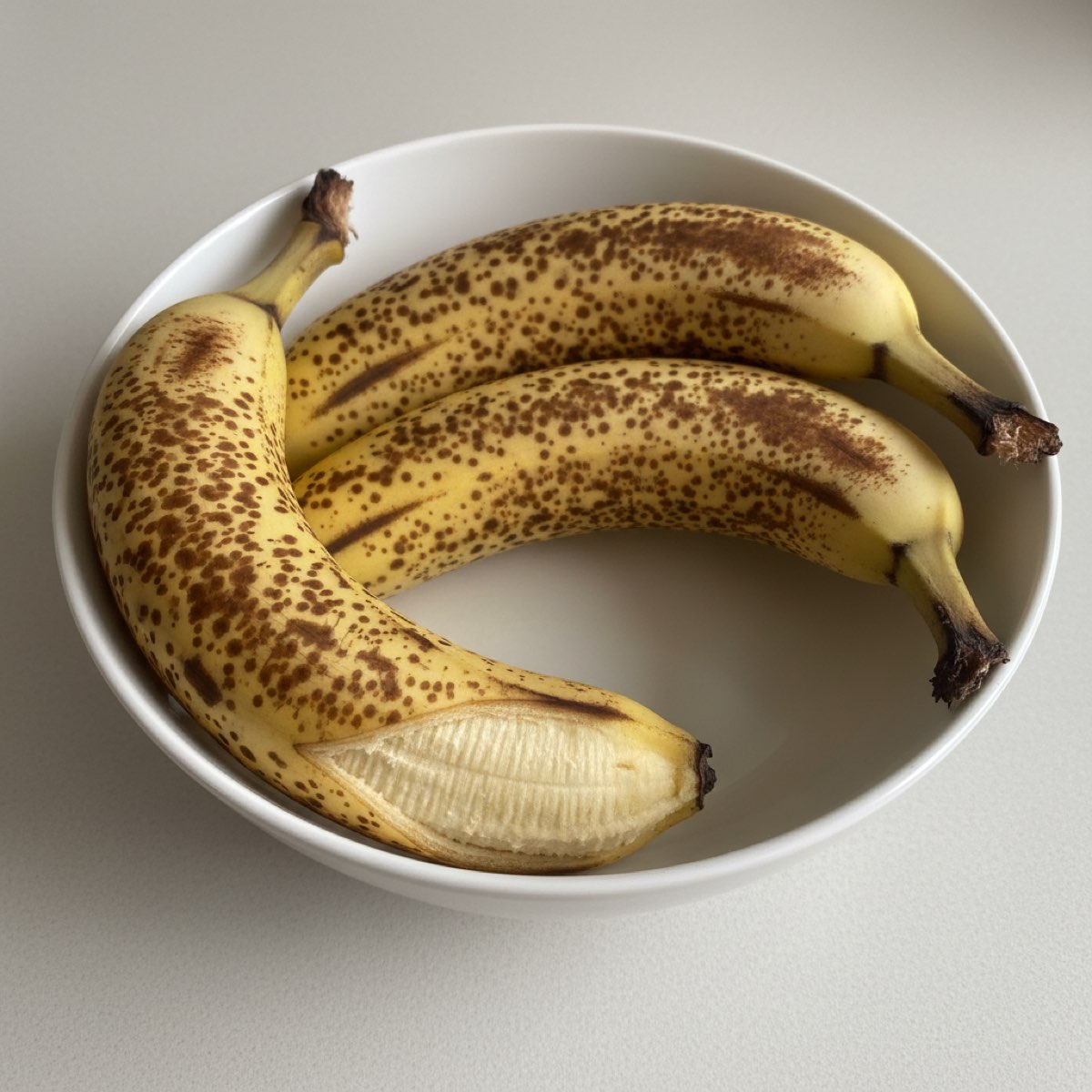 Three heavily speckled, overripe bananas in a white bowl, one with its skin split open.